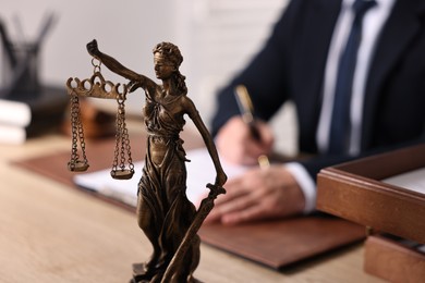 Photo of Notary writing notes at wooden table in office, focus on statue of Lady Justice