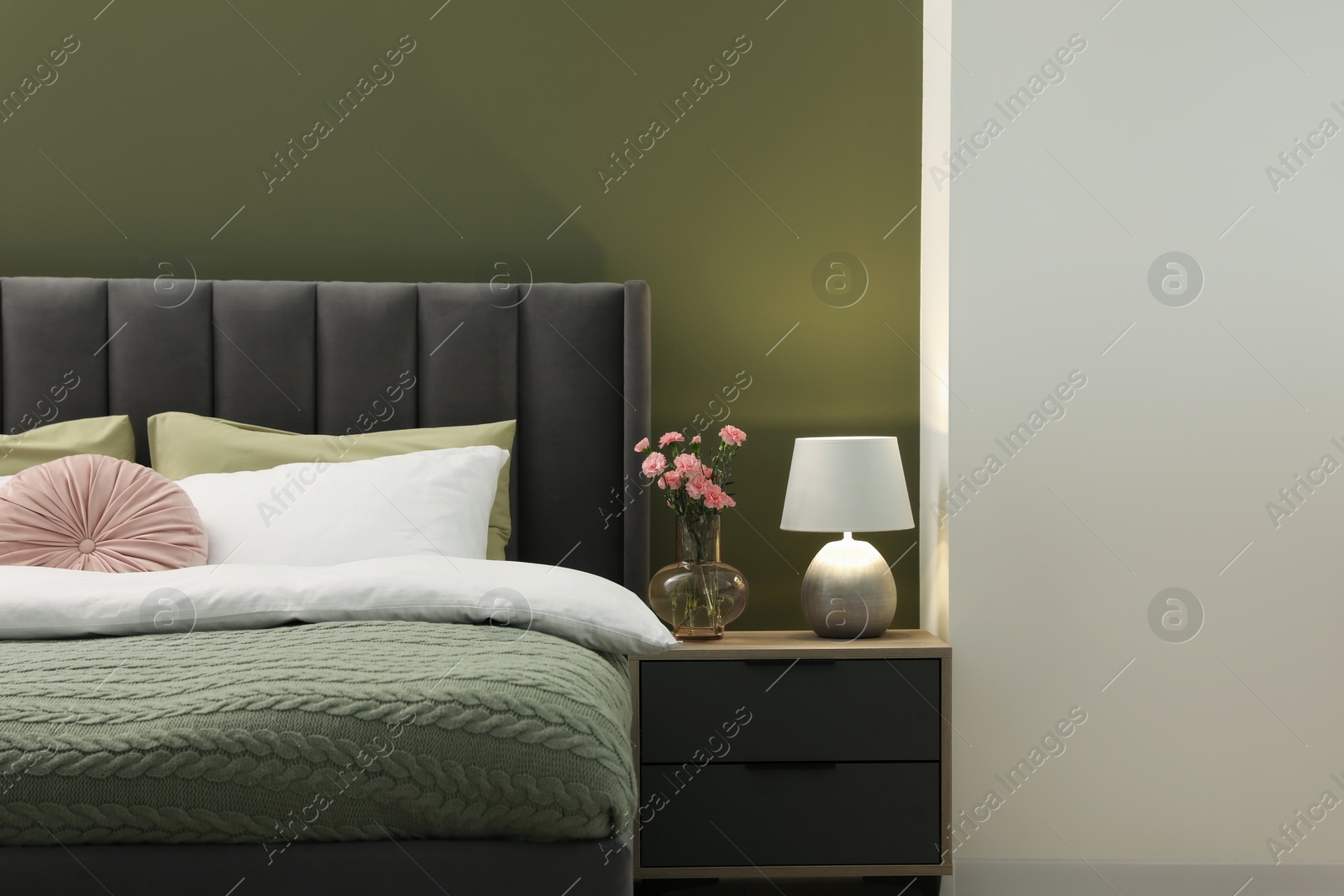 Photo of Large comfortable bed, nightstand, lamp and beautiful flowers in stylish room. Interior design