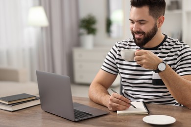 Young man with cup of coffee watching webinar at table in room