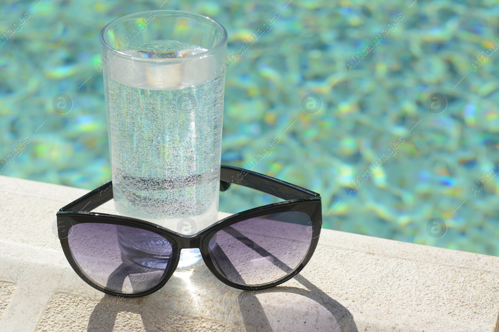 Photo of Stylish sunglasses and glass of water near outdoor swimming pool on sunny day, closeup