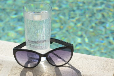 Photo of Stylish sunglasses and glass of water near outdoor swimming pool on sunny day, closeup