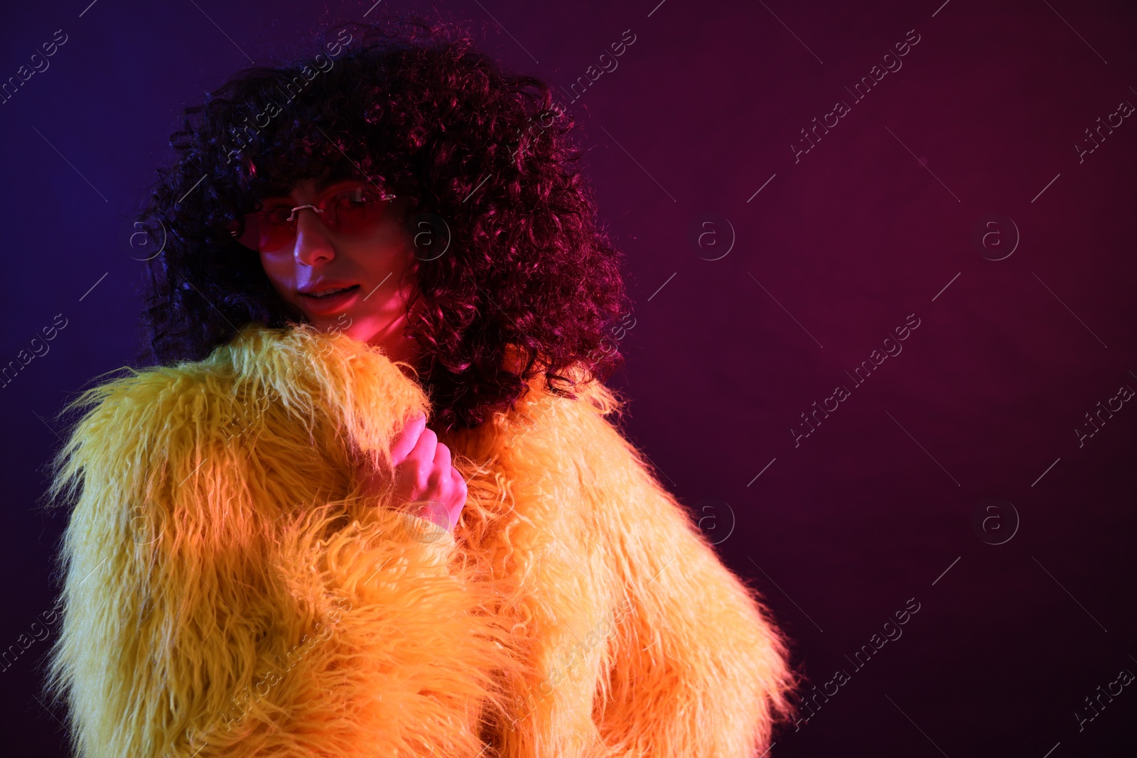 Photo of Beautiful young woman in yellow fur coat and sunglasses on color background in neon lights. Space for text