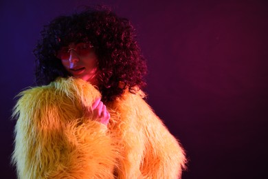 Photo of Beautiful young woman in yellow fur coat and sunglasses on color background in neon lights. Space for text