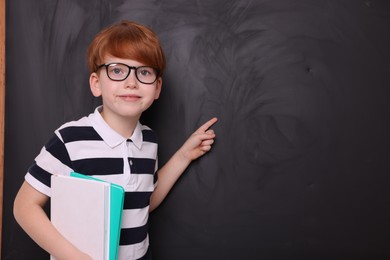 Cute schoolboy in glasses with books pointing at something on blackboard. Space for text