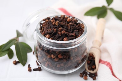 Photo of Aromatic cloves in glass jar, scoop and green leaves on white table, closeup