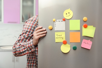 Photo of Man opening refrigerator door with paper sheets and magnets at home, closeup