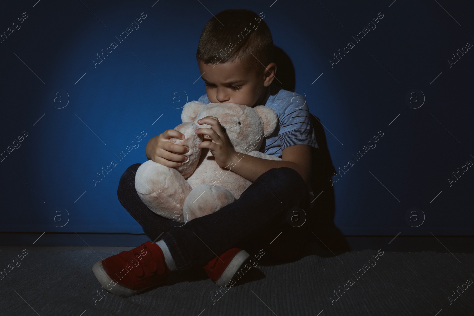 Photo of Sad little boy with teddy bear near blue wall. Domestic violence concept