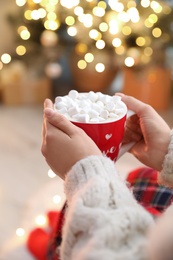 Photo of Woman holding cup of hot drink with marshmallows indoors, closeup. Magic Christmas atmosphere