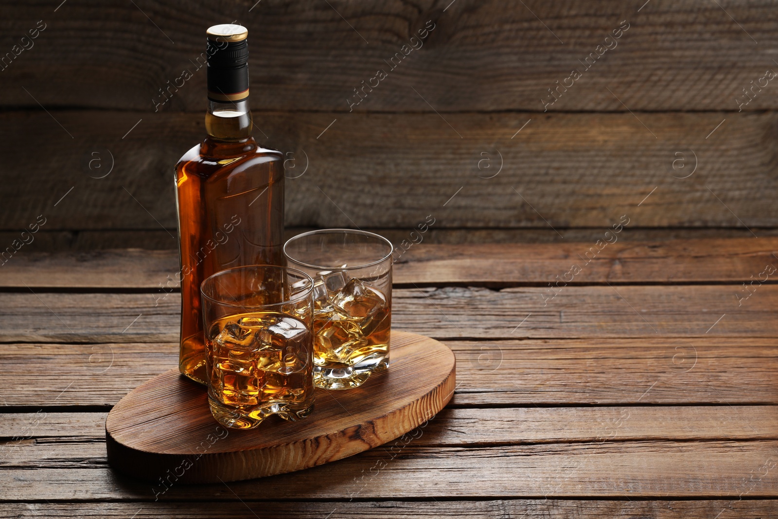 Photo of Whiskey with ice cubes in glasses and bottle on wooden table, space for text