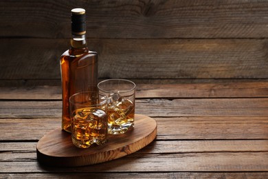 Whiskey with ice cubes in glasses and bottle on wooden table, space for text