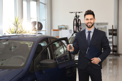 Photo of Young salesman with key and clipboard near car in dealership