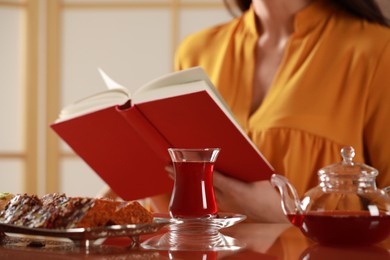 Photo of Woman reading book, glass of traditional Turkish tea and snacks on table indoors, selective focus