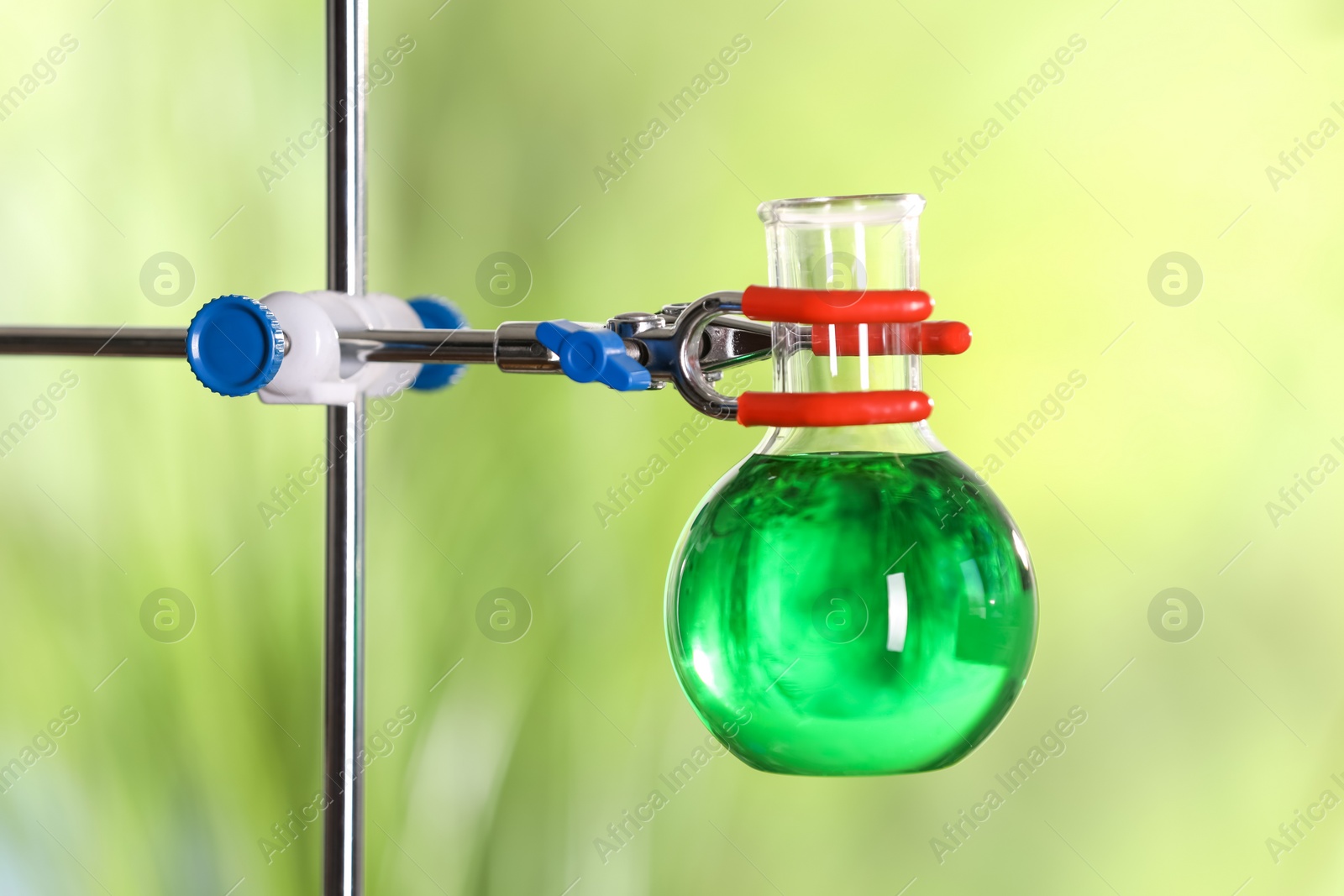 Photo of Flask with green liquid on retort stand against blurred background, closeup