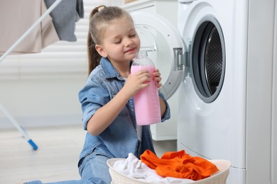 Photo of Little girl smelling fabric softener near washing machine and basket with dirty clothes in bathroom, space for text