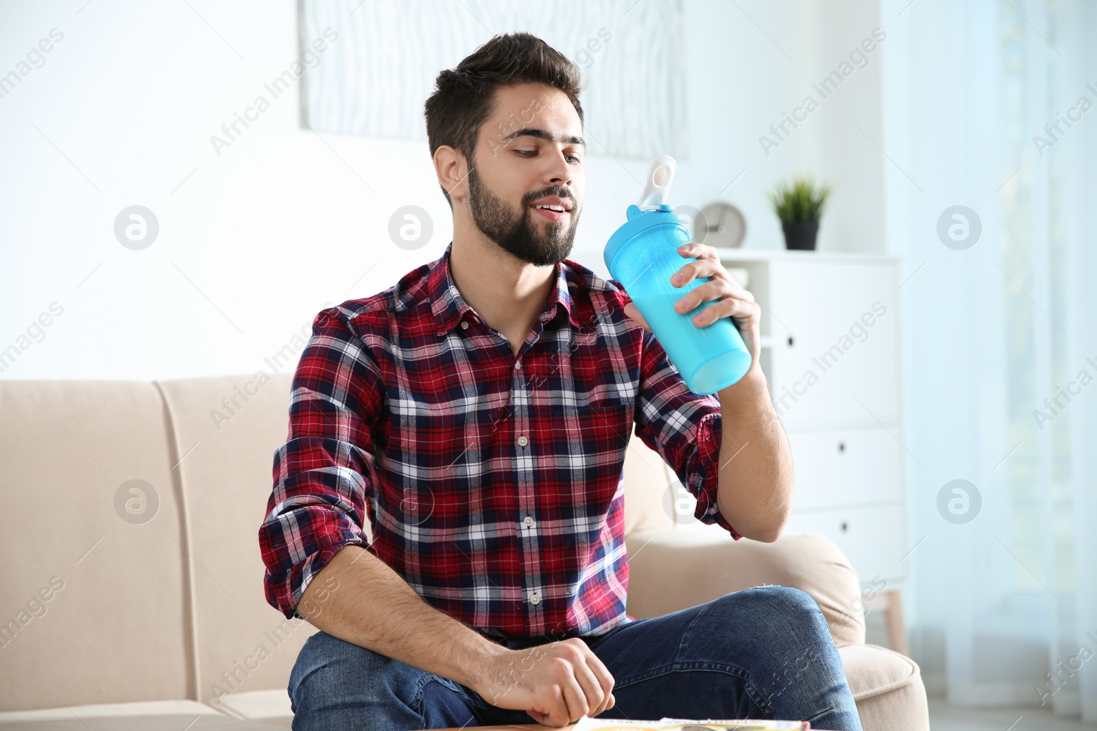 Photo of Young man with bottle of protein shake sitting on sofa at home