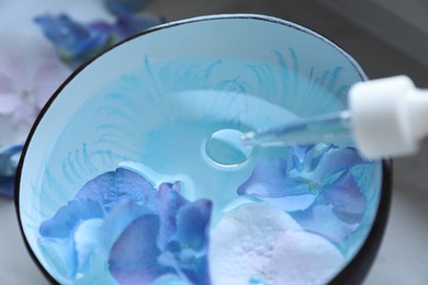 Photo of Dripping essential oil from pipette into bowl with water and flowers, closeup