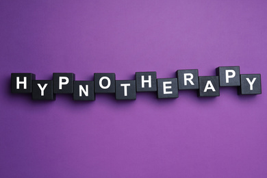 Photo of Black wooden blocks with word HYPNOTHERAPY on purple background, flat lay