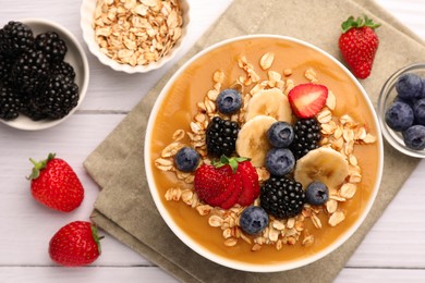 Photo of Delicious smoothie bowl with fresh berries, banana and oatmeal on white wooden table, flat lay