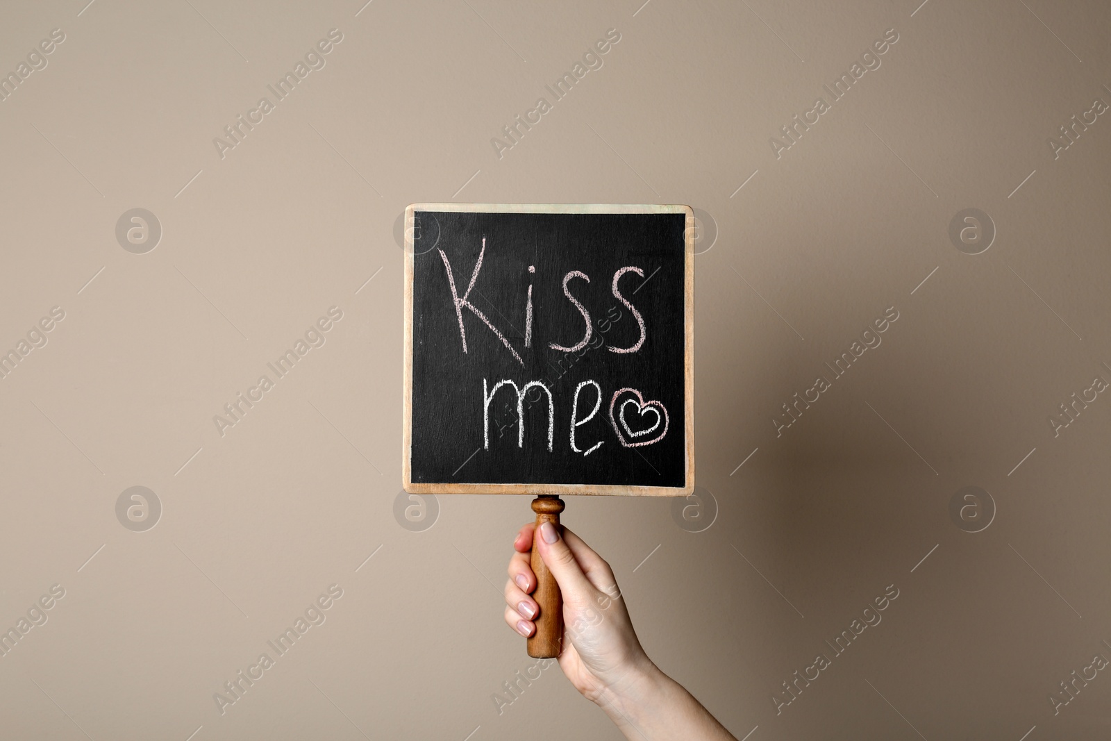 Photo of Woman holding small chalkboard with phrase Kiss Me on beige background, closeup