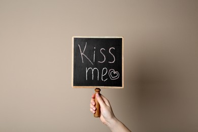 Woman holding small chalkboard with phrase Kiss Me on beige background, closeup