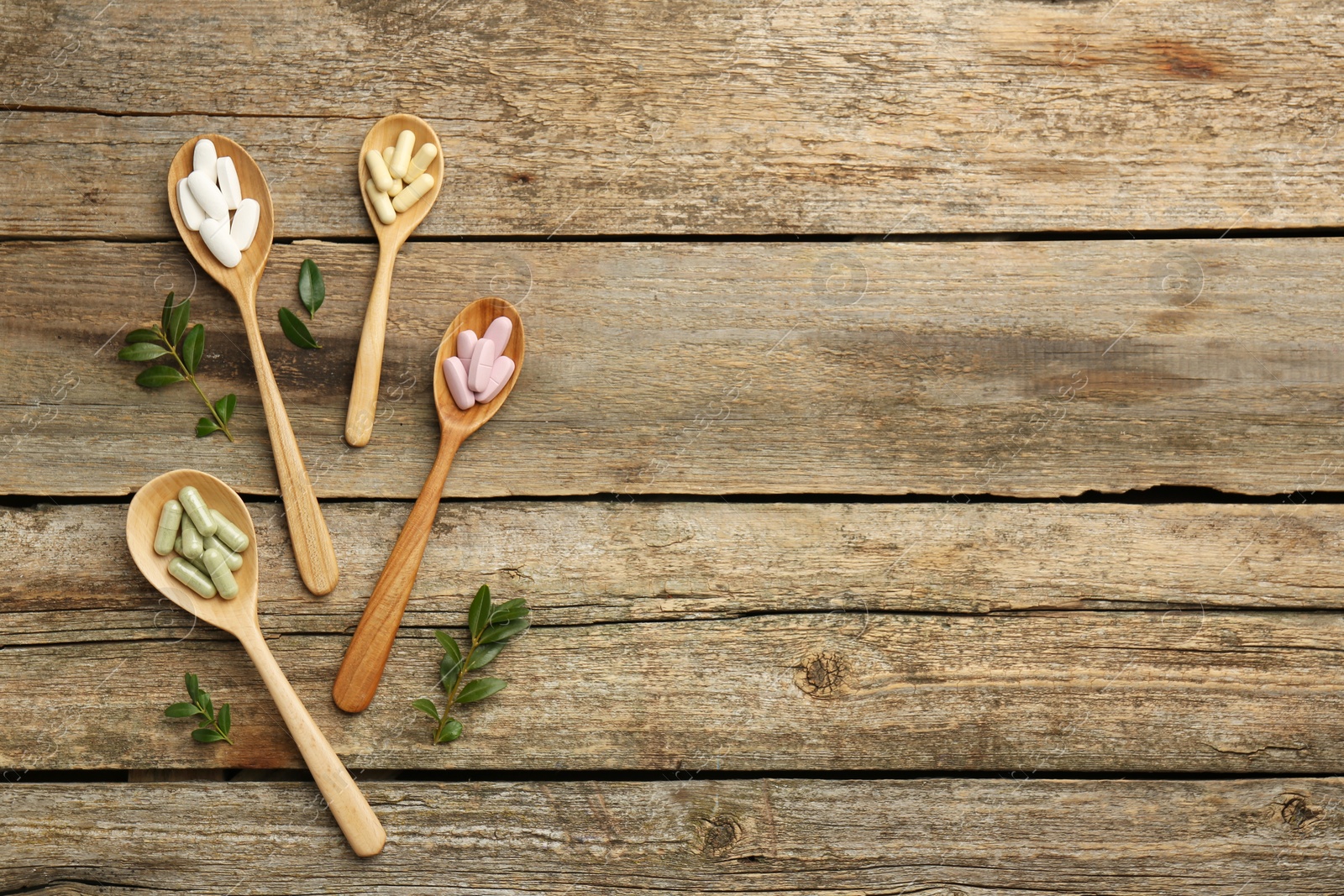 Photo of Vitamin capsules in spoons and leaves on wooden table, flat lay. Space for text