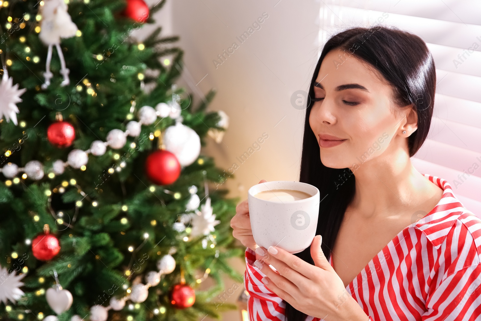 Photo of Young woman with cup of coffee near Christmas tree at home, space for text