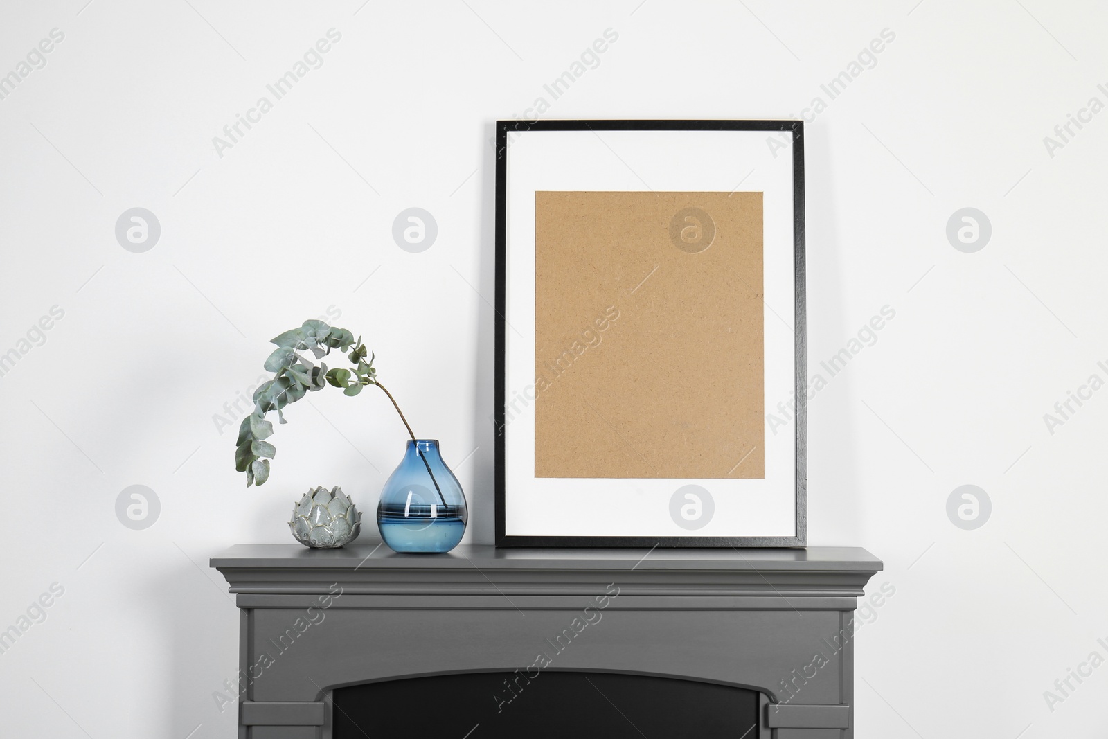 Photo of Empty frame and eucalyptus branch in vase on fireplace near white wall indoors. Interior design