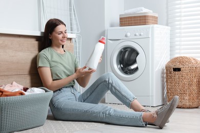 Photo of Woman sitting on floor near washing machine and holding fabric softener in bathroom