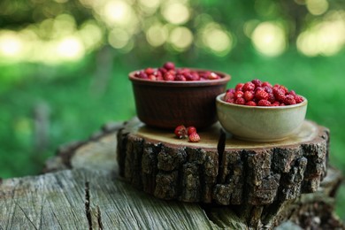 Bowls of tasty wild strawberries on stump against blurred background, closeup. Space for text