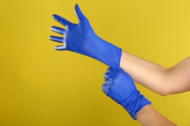 Woman putting on blue latex gloves against yellow background, closeup of hands