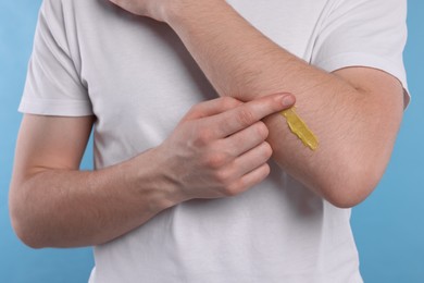 Man applying yellow ointment onto his arm on light blue background, closeup