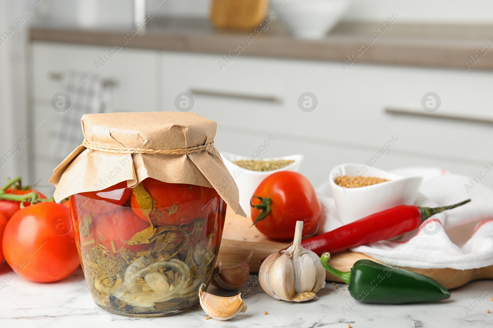 Photo of Pickled tomatoes in glass jar and products on white marble table in kitchen