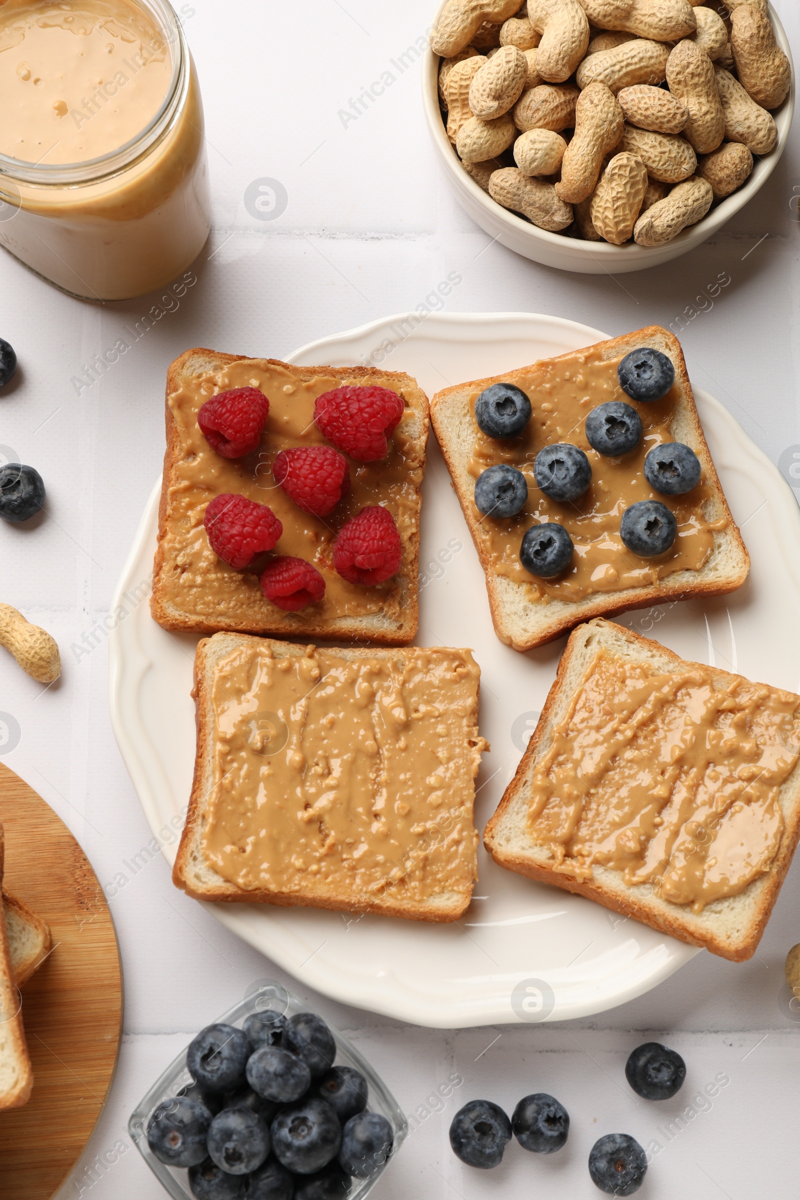 Photo of Delicious toasts with peanut butter, raspberries, blueberries and nuts on white tiled table, flat lay