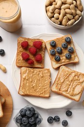 Photo of Delicious toasts with peanut butter, raspberries, blueberries and nuts on white tiled table, flat lay