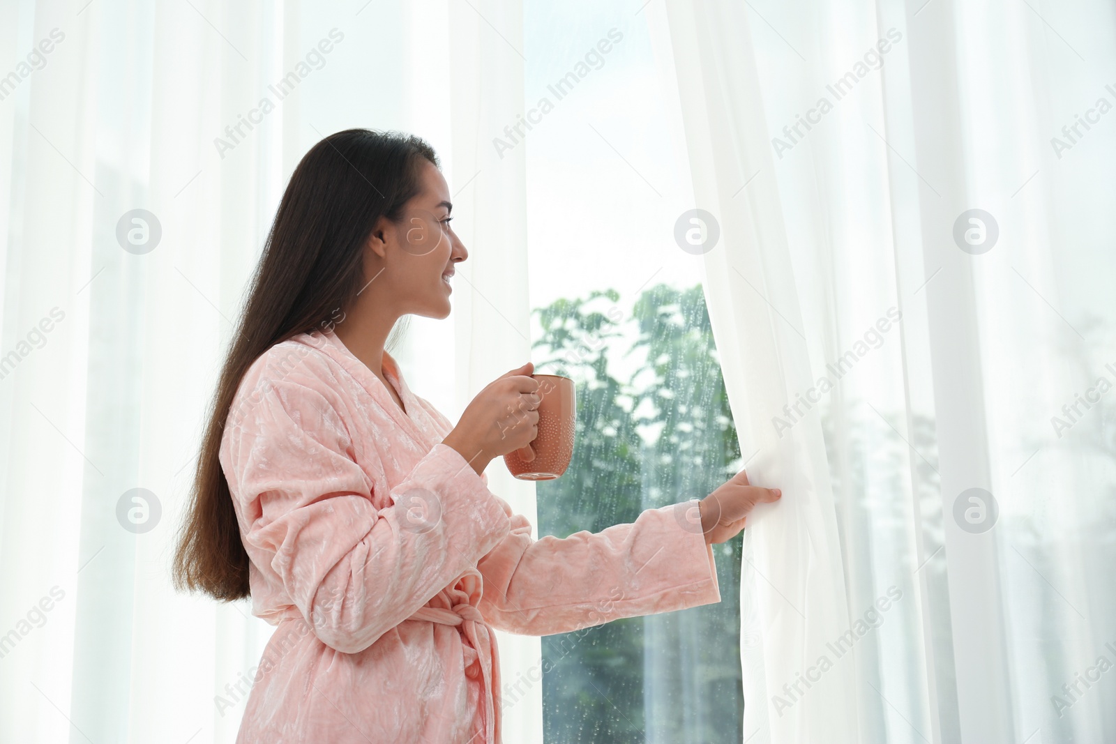 Image of Young woman with cup of coffee opening curtains at home. Good morning