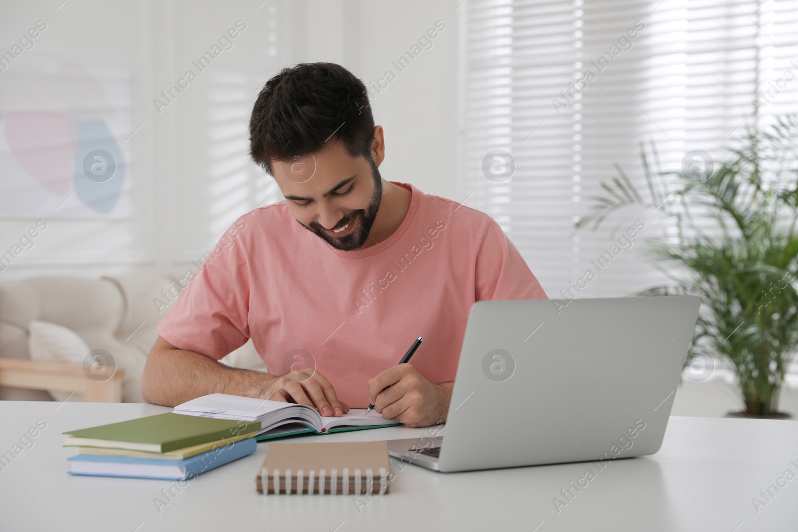 Photo of Young man writing down notes during webinar at table in room