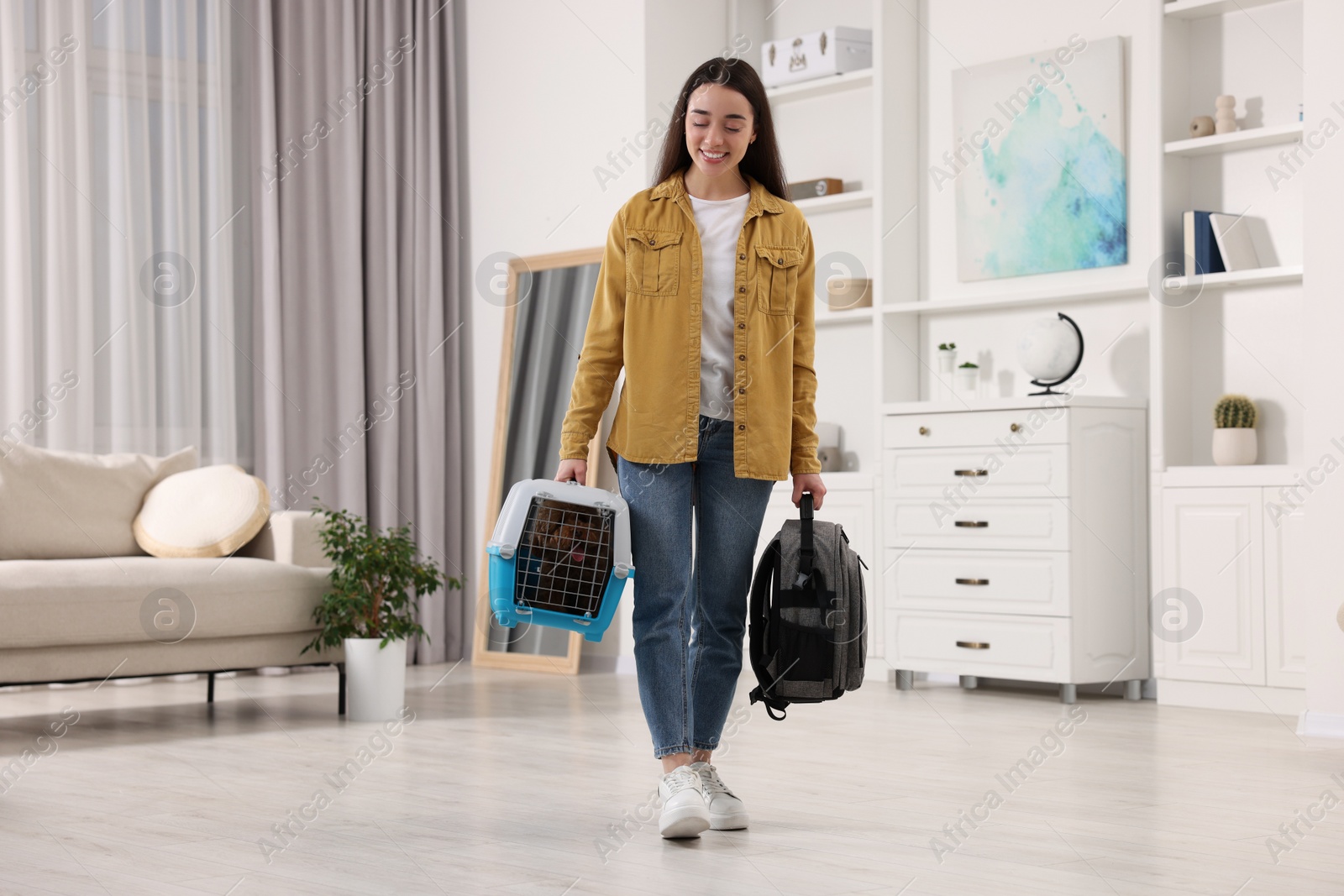 Photo of Travel with pet. Smiling woman holding carrier with dog and backpack at home