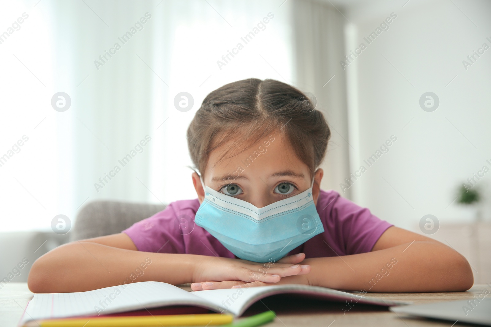 Photo of Distance learning, online studying at home due to Covid-19 pandemic. Sad girl in protective mask doing homework in room