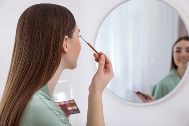 Photo of Beautiful young woman applying eyeshadow with brush near mirror indoors