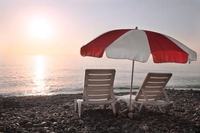 Empty sunbeds and umbrella on stone beach near sea at sunset