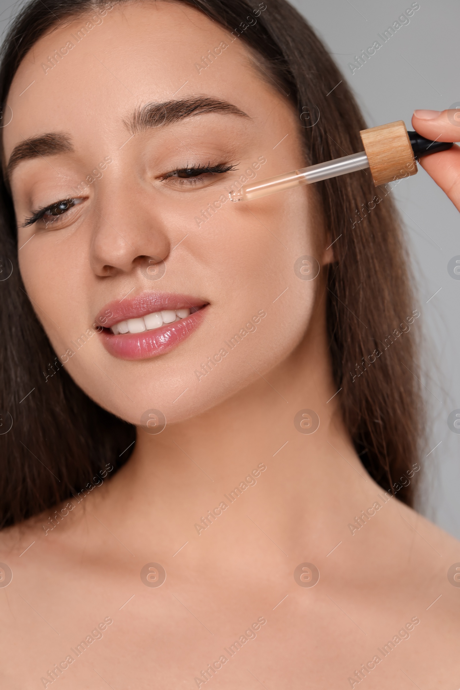 Photo of Beautiful young woman applying essential oil onto face on grey background, closeup