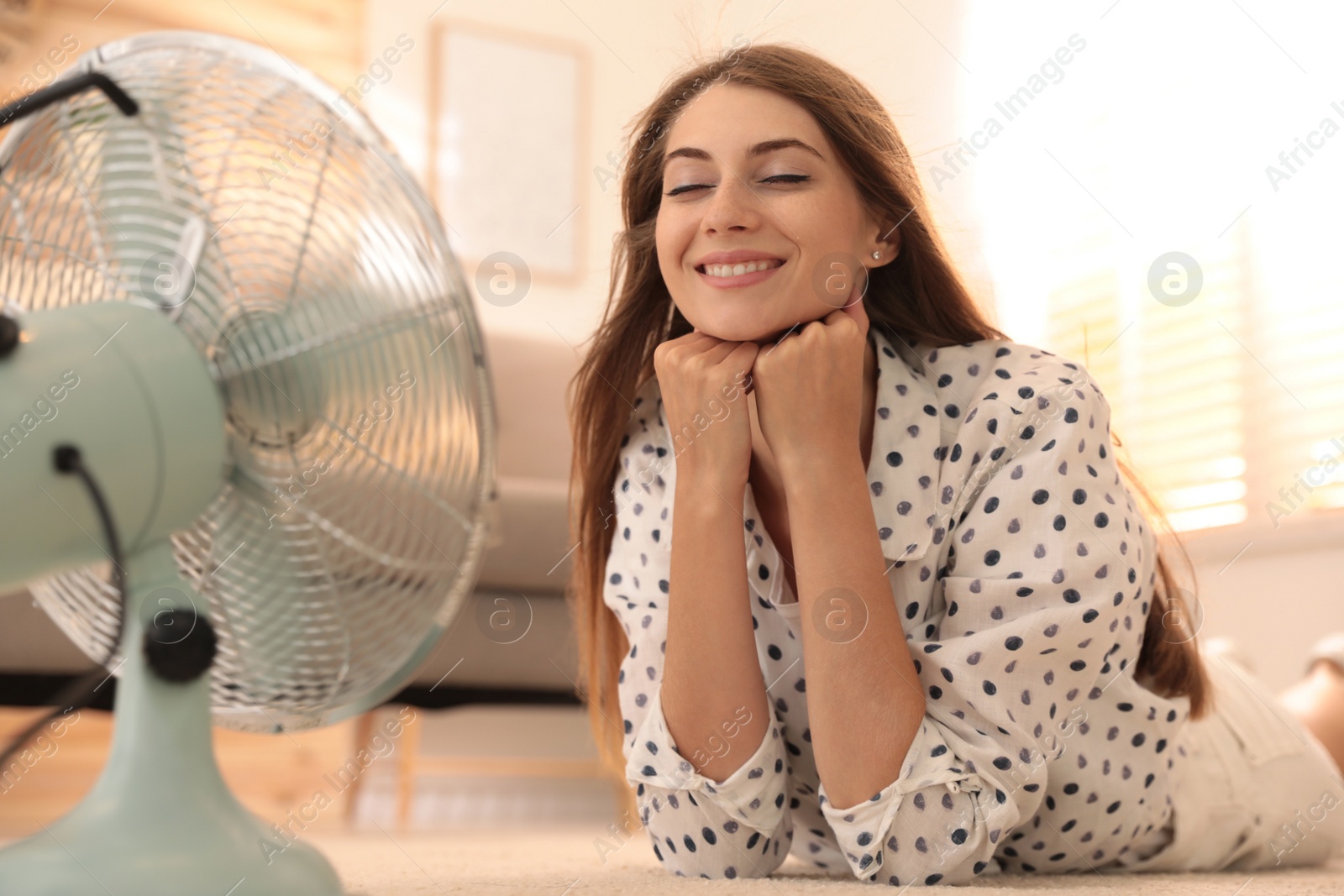 Photo of Woman enjoying air flow from fan on floor in living room. Summer heat