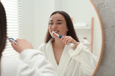 Photo of Young woman brushing her teeth with electric toothbrush near mirror in bathroom