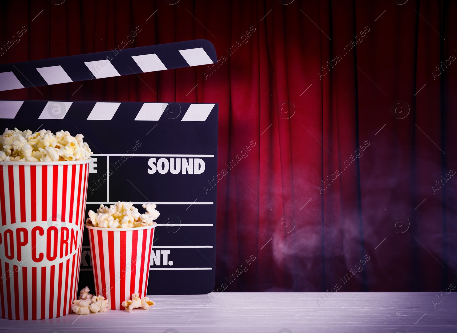 Image of Delicious popcorn and clapperboard on wooden table against closed red main curtain, space for text