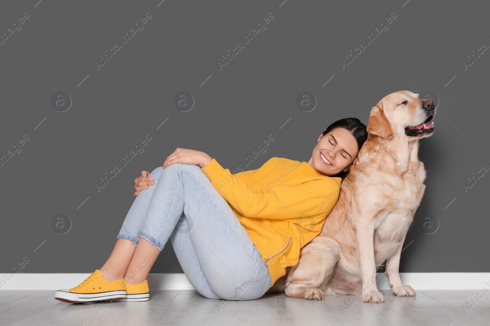 Photo of Happy woman with cute Labrador Retriever on floor against grey wall