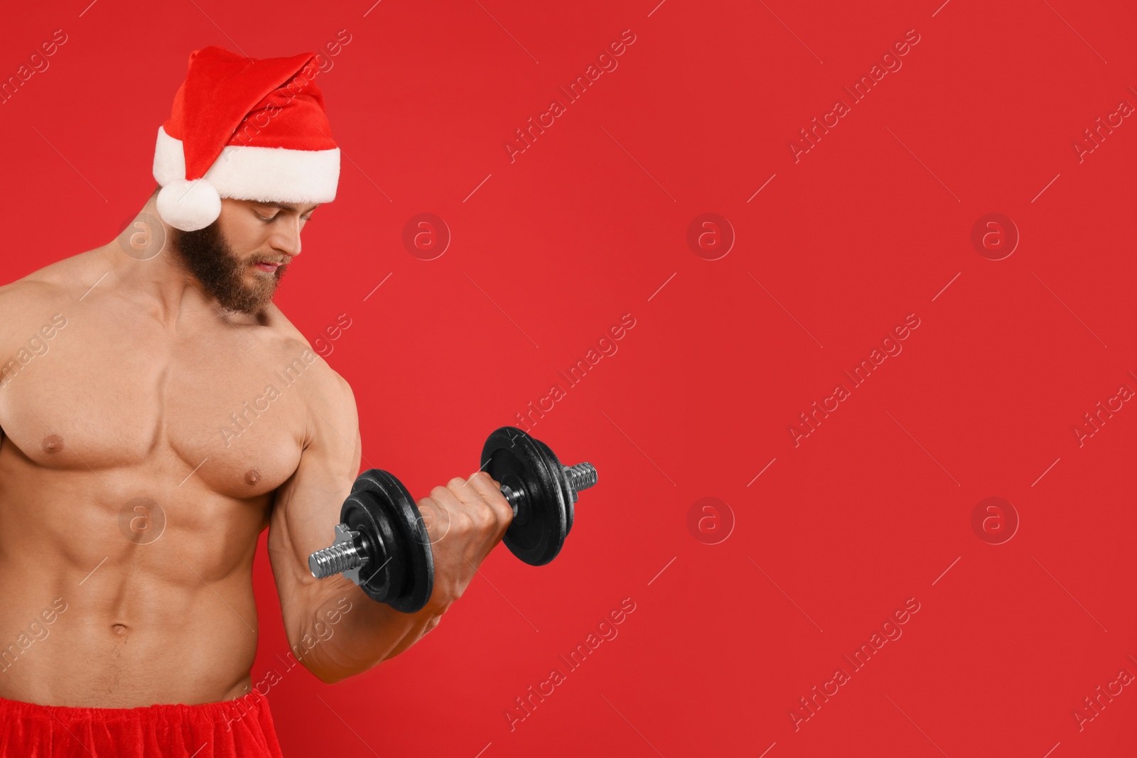 Photo of Muscular young man in Santa hat with dumbbell on red background, space for text