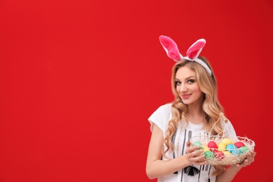 Photo of Beautiful young woman in bunny ears headband holding basket with Easter eggs on color background, space for text