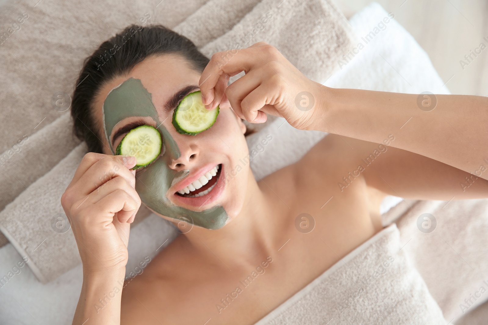 Photo of Pretty woman with clay mask on her face holding cucumber slices in spa salon, above view