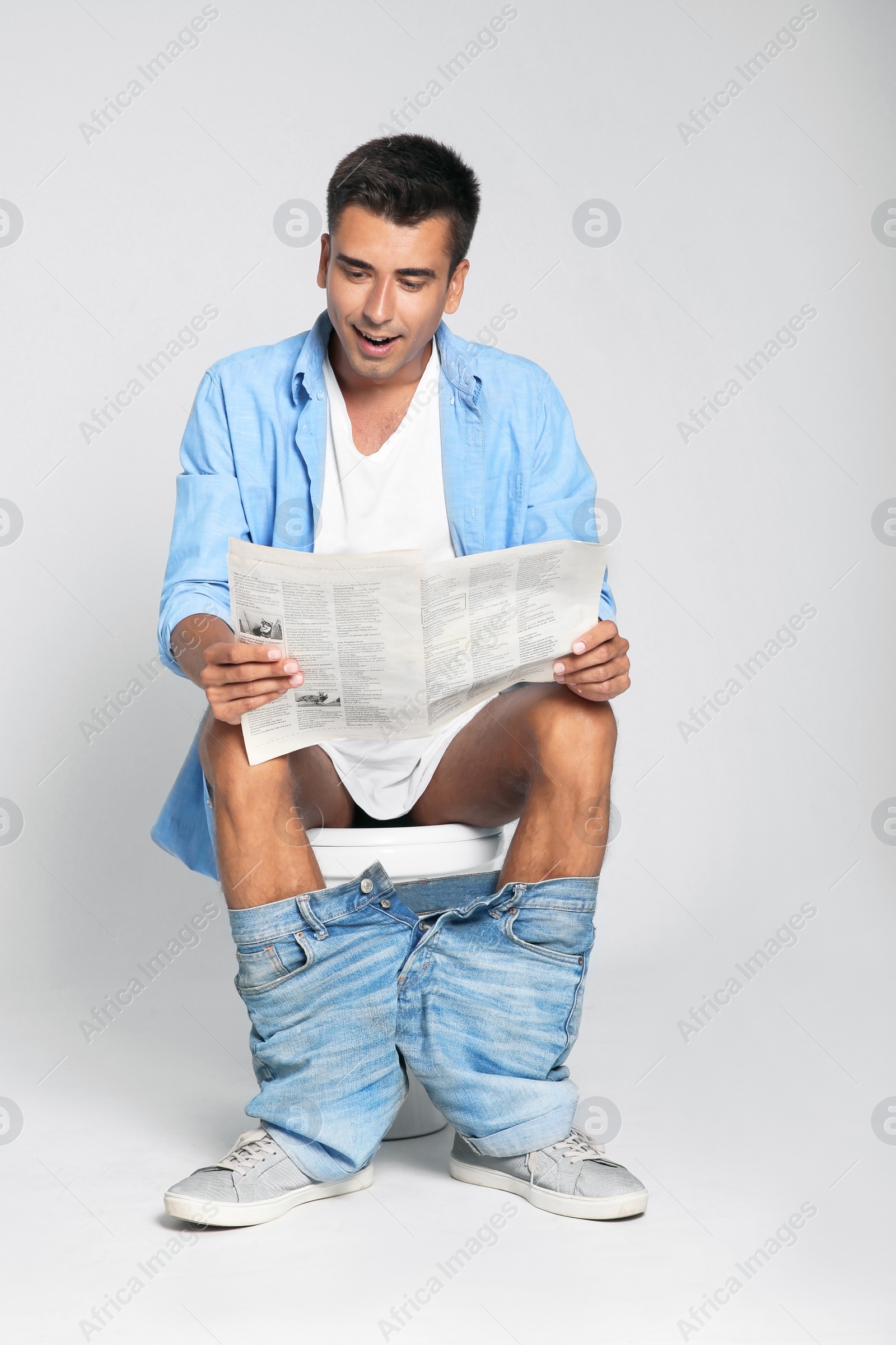 Photo of Young man reading newspaper while sitting on toilet bowl against gray background