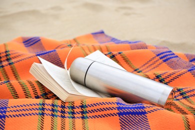 Metallic thermos with hot drink, open book and plaid on sandy beach, closeup